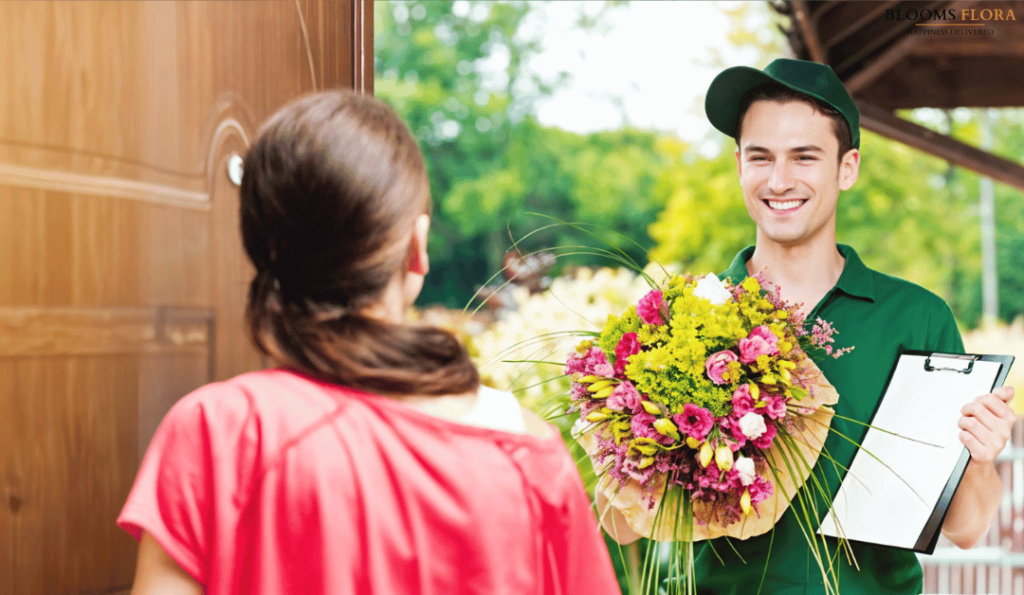 A delivery person in a green uniform smiles as they hand a vibrant flower bouquet and clipboard to a woman at her doorstep.